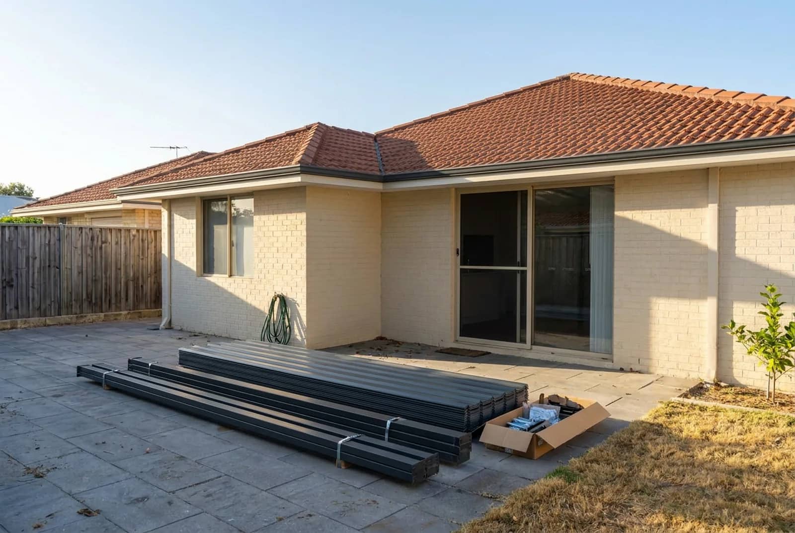 A flat-packed steel patio kit laid out on the paving behind a Perth home, beams and posts strapped in bundles beside a box of brackets and bolts